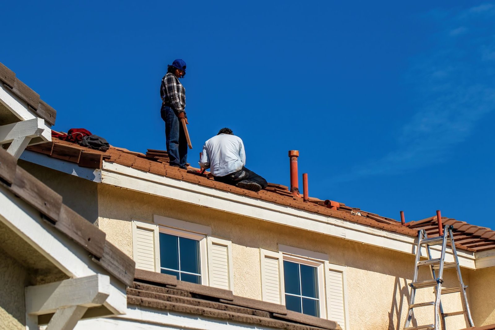 Two roofers laying clay tiles on a house under a blue sky
